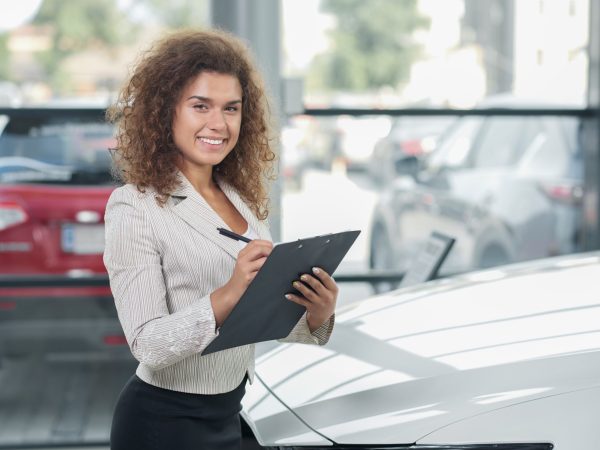 Female manager of car dealership standing in showroom and posing. Pretty woman with curly hair smiling, looking at camera. Car dealer holding black folder and pen.